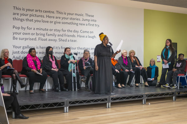 A woman reads at a microphone, backed by a seated group of women on stage with a background wall text reading 'This is your arts centre'.