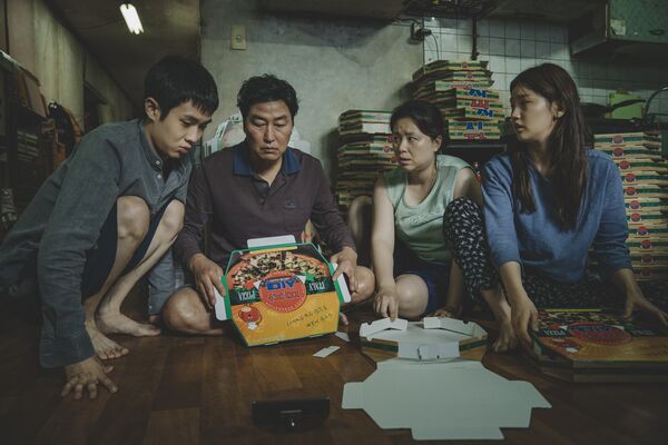 A family sit together on the floor of a kitchen surrounded by pizza boxes. 