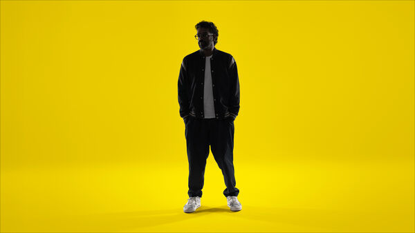 Romesh Ranganthan with his hands in pockets - on a yellow background