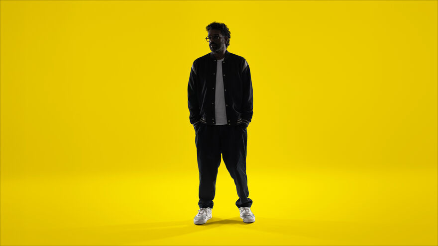 Romesh Ranganthan with his hands in pockets - on a yellow background