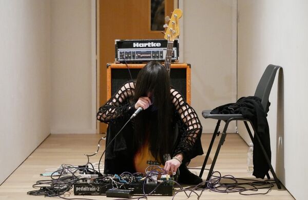 Person with microphone to mouth while looking down surrounded by cables. Guitar, amp and mixer behind