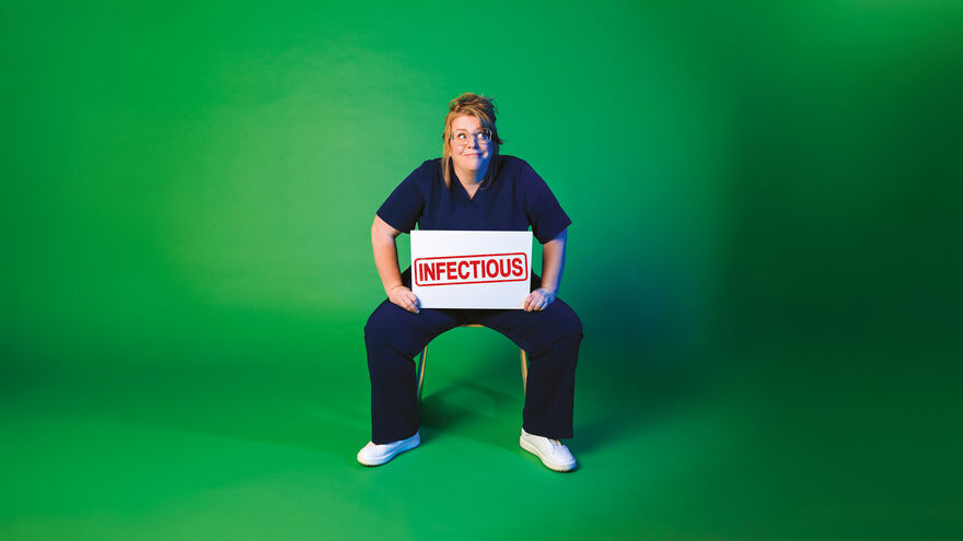 Georgie sits on a chair in nurses scrubs in front of a green background holding a sign with the word Infectious on
