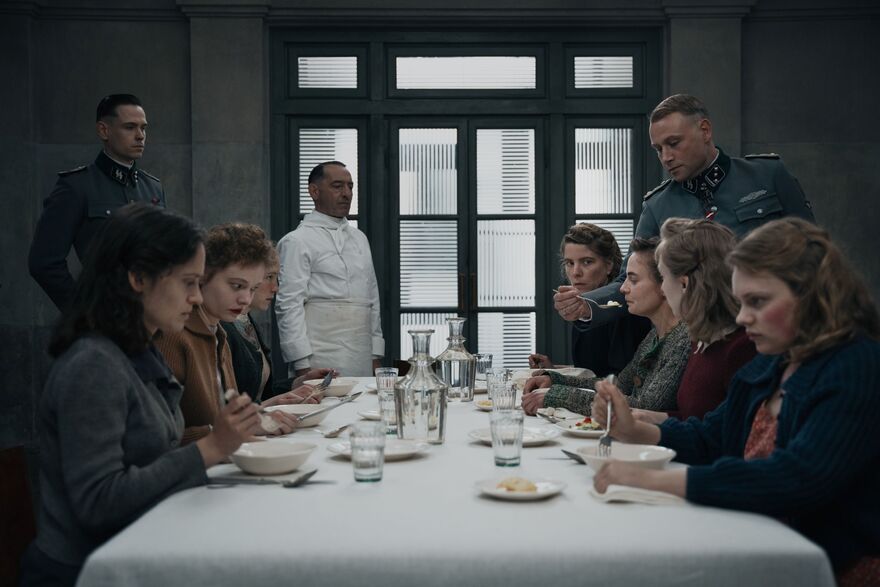 A group of women sit around a dining table watched over by two Nazi officers. One woman is being spoon fed. 