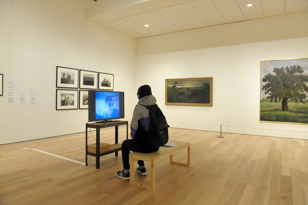 Person sitting down in front of video on tv screen in exhibition with art on walls. 