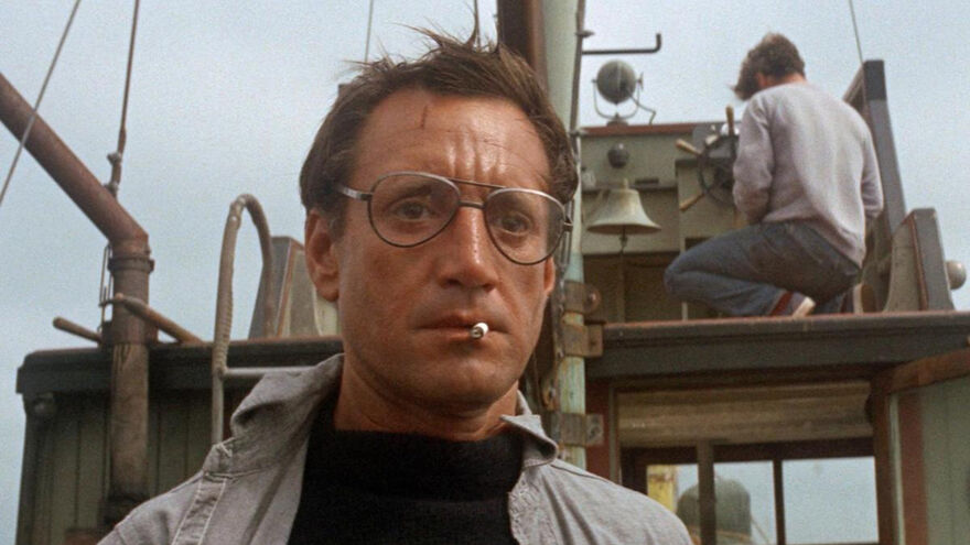 A man, wearing glasses, looks concerned with a cigarette between his lips. He stands on the deck of a trawler.  