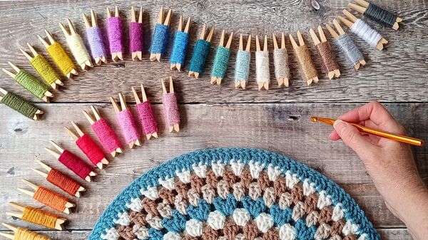 A variety of colourful yarn wrapped around pegs, a blue crochet cushion and a hand holding a crochet hook
