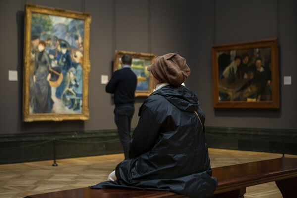A person sits in hat and coat on a bench in a gallery looking at the artwork.