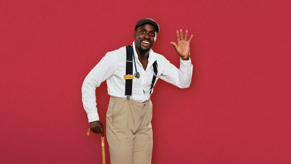 Michael Akadiri in biege trousers, white shirt and braces waving and holding a walking stick