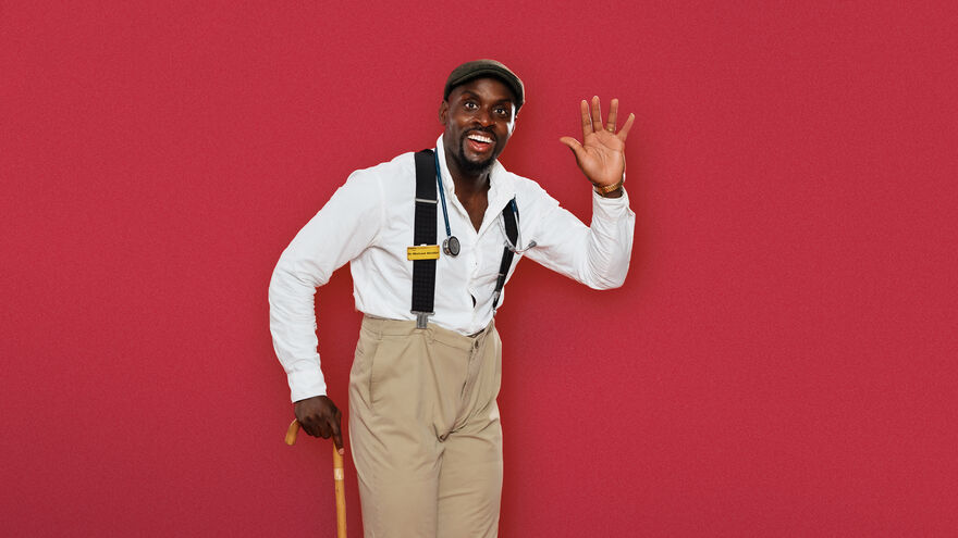 Michael Akadiri in biege trousers, white shirt and braces waving and holding a walking stick