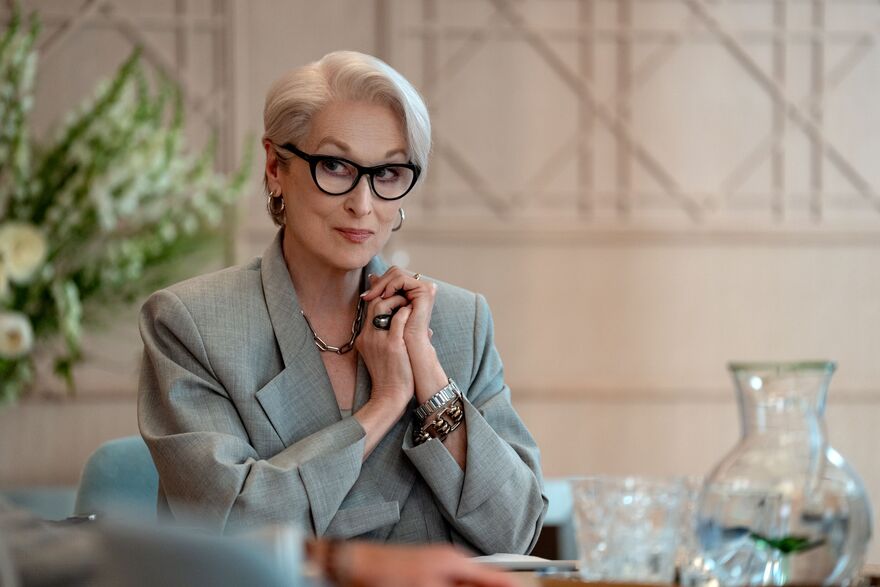 An older woman sits at a table with her hands together, she is looking to the side. 
