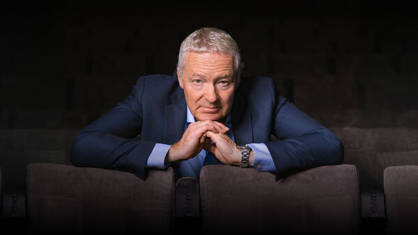 Rory Bremner is ablue suit with his hands togther leaning over some chairs 