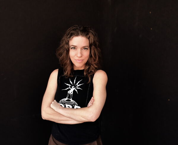 Ani DiFranco stood with her arms folded in a balck shirt with a black background