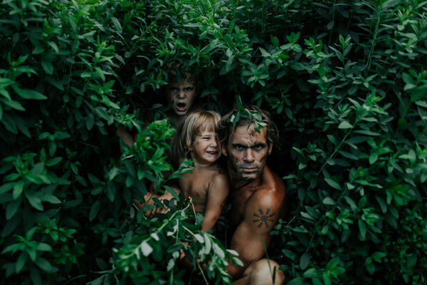 A father and two children peer out from a green bush. 