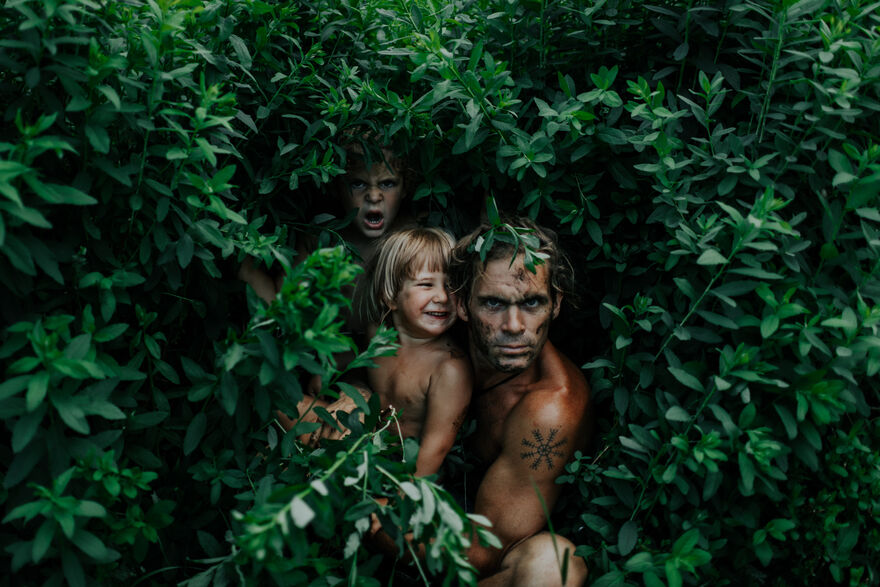 A father and two children peer out from a green bush.