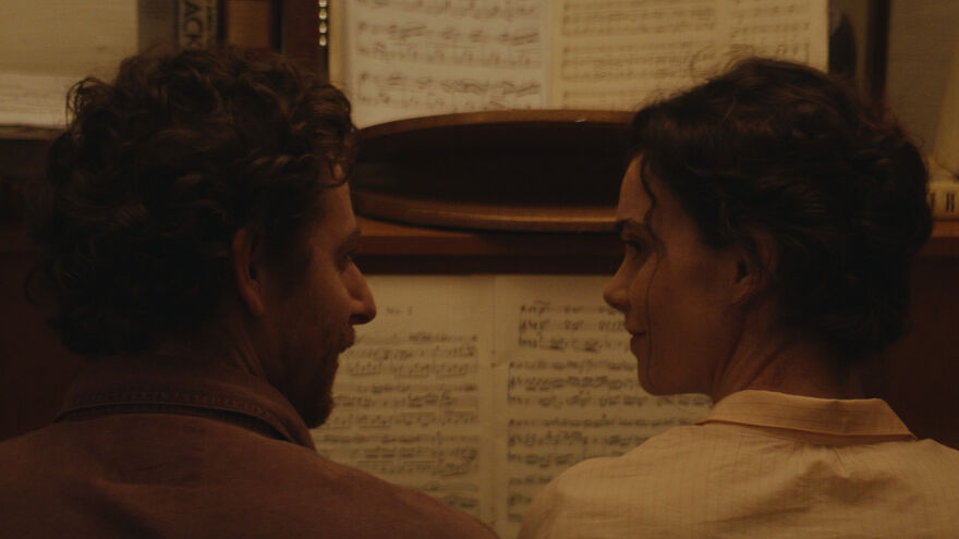 A man and woman sit side by side at a piano gazing at one another. 