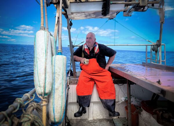 A fisherman sits on a boat in bright orange waders drinking a hot drink. 