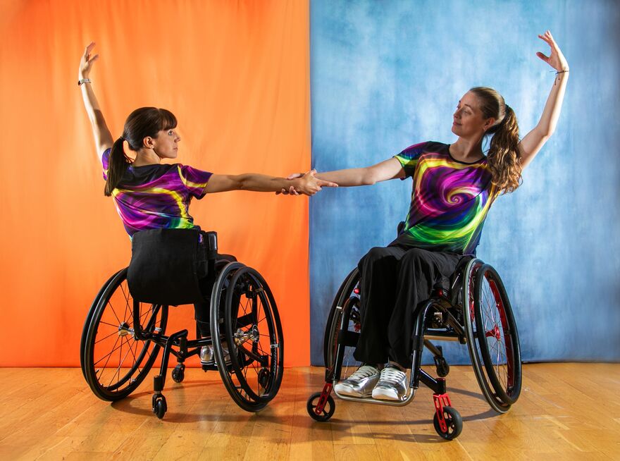 Two women in wheelchairs holding arms with their other arm in the air, background is split in half with one side orange and the other blue