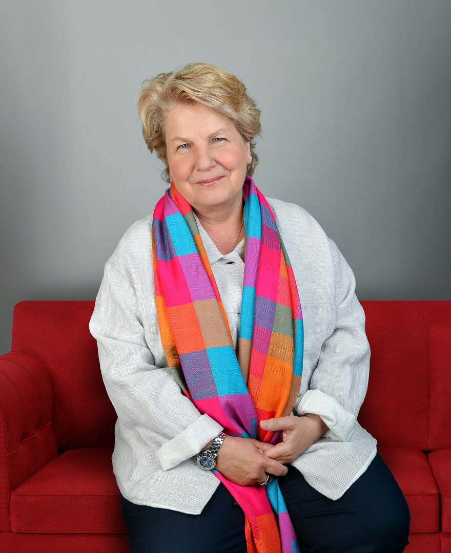 Sandi Toksvig sitting on a red sofa wearing a long pink chequered scarf
