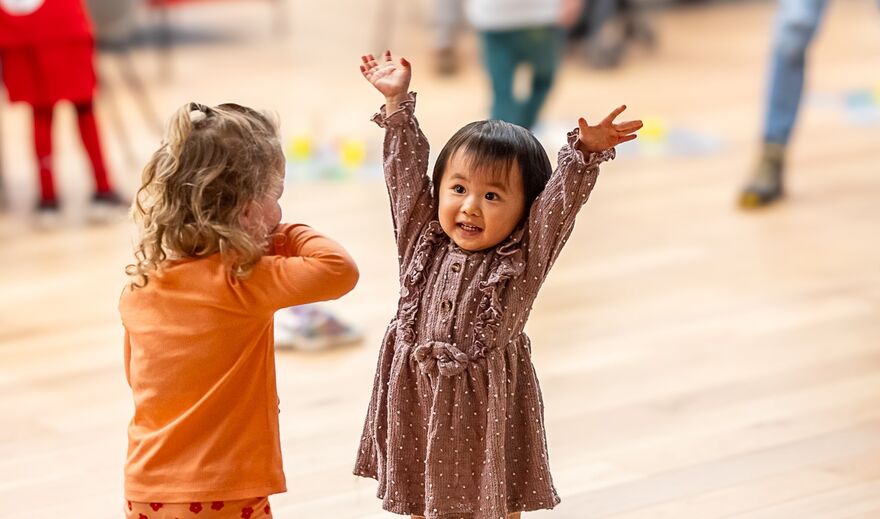 Two young children, one has hands in the air the other covering their mouths