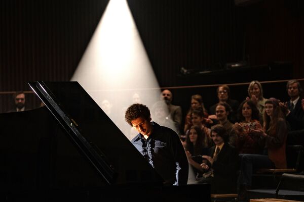 A man sits at a piano, behind him an audience applauds. 