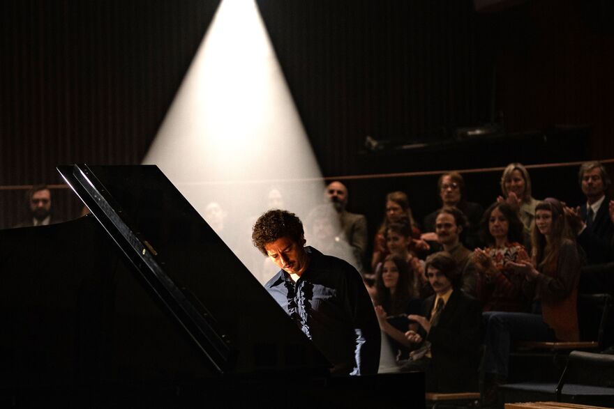 A man sits at a piano, behind him an audience applauds. 