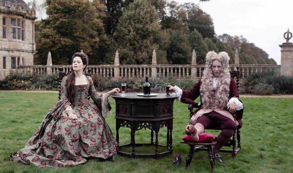 A couple in 18th century dress sit in the grounds of an expansive estate. A couple in 18th century dress sit in the grounds of an expansive estate.