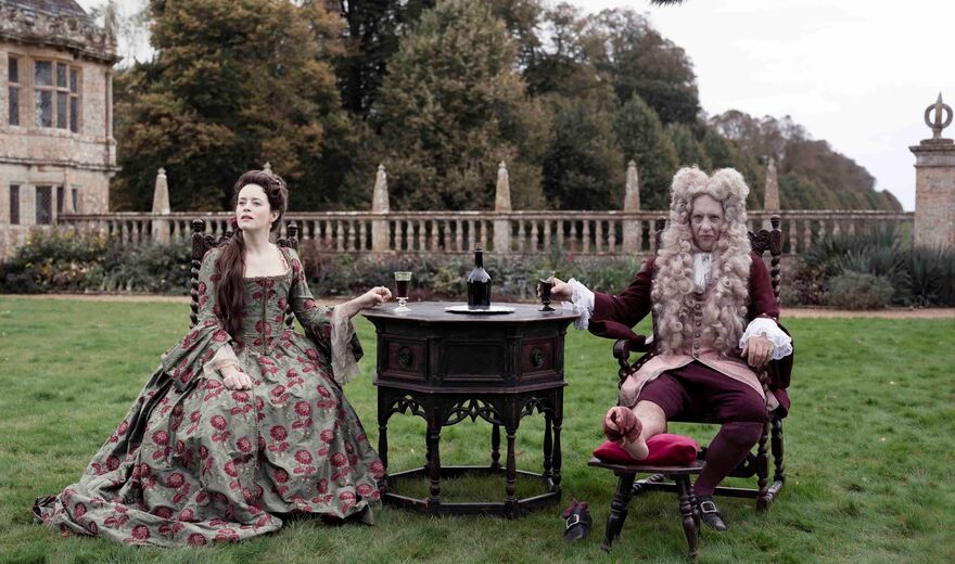 A couple in 18th century dress sit in the grounds of an expansive estate. 