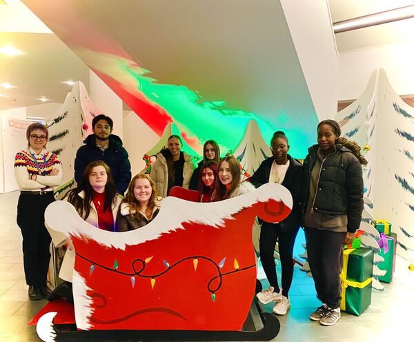 A group of young people pose for a festive photo around a large red Santa's sleigh prop, surrounded by white Christmas tree cutouts and wrapped gifts.