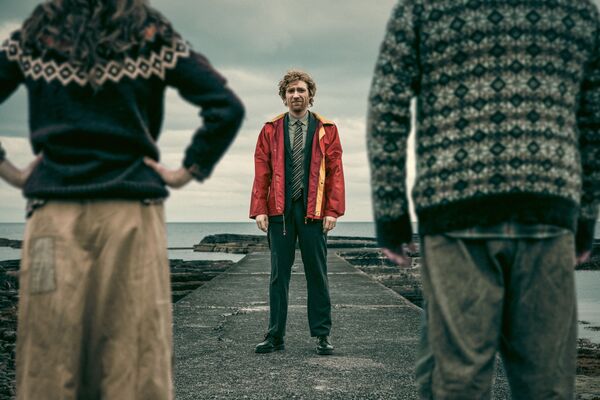A man, wearing a suit and red raincoat, stands on a pier facing two people in jumpers. They have their backs to camera. The sea is in the background. 