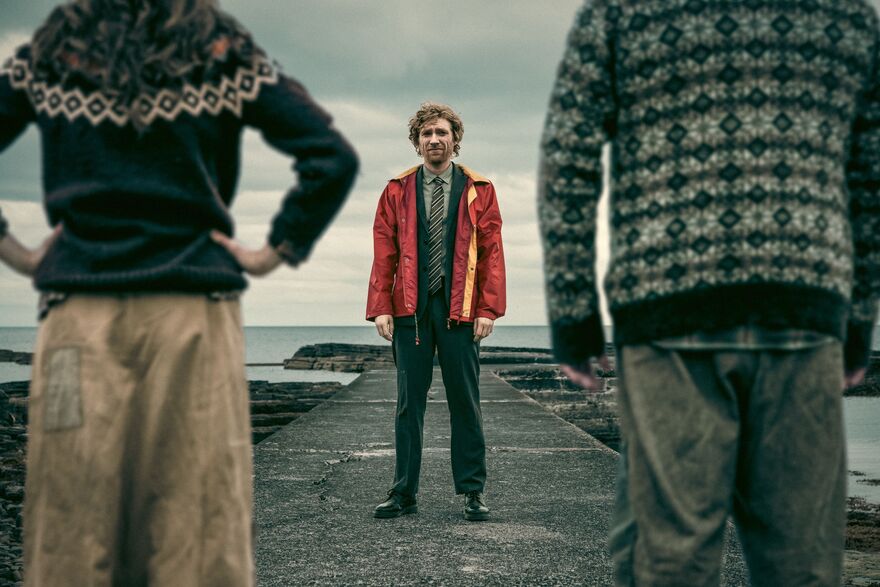 A man, wearing a suit and red raincoat, stands on a pier facing two people in jumpers. They have their backs to camera. The sea is in the background. 