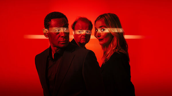 David Harewood, Toby Jones and Caitlin Fitzgerald against a red background. Their eyes are highlighted with a strip of bright light. 