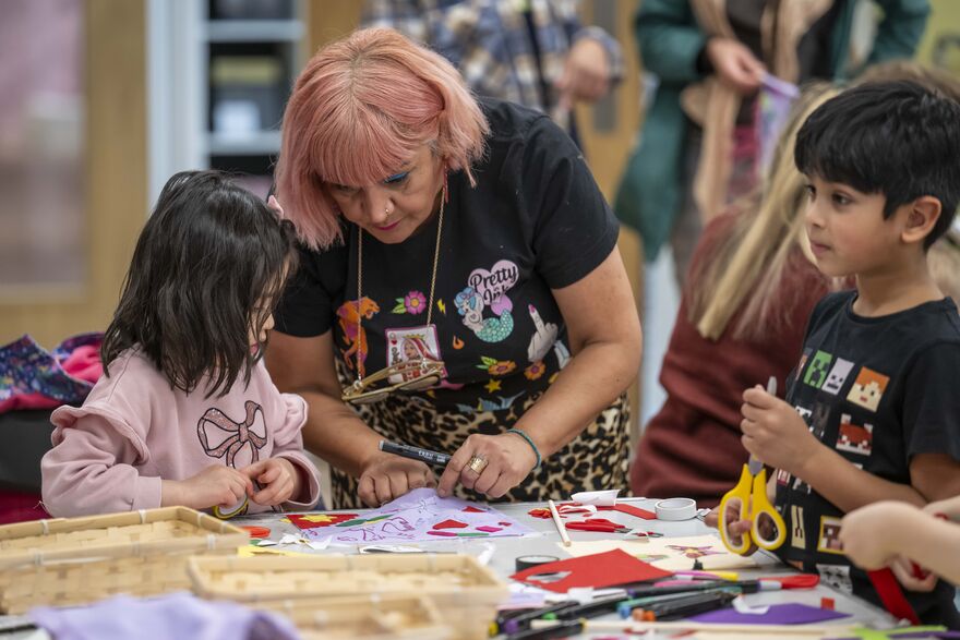 a lady helps a young girl with an activity as a young boy stands nearby holding scissors