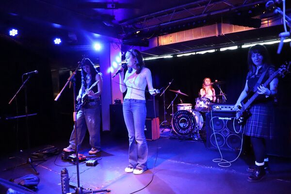 The all-female alternative band Astraea performing live on a stage with blue lighting, featuring a lead singer, guitarist, bassist, and a drummer with a Warwick Bandsoc drum skin. The all-female alternative band Astraea performing live on a stage with blue lighting, featuring a lead singer, guitarist, bassist, and a drummer with a Warwick Bandsoc drum skin.
