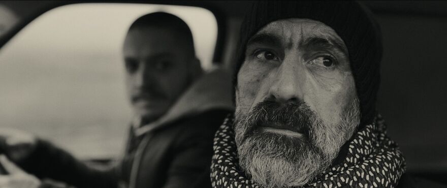 A back and white image of an older man looking out the window on the passenger side of a car. A young man can be seen in the background behind the wheel. 