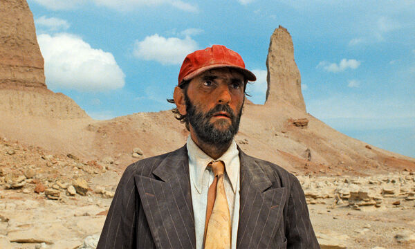 A bearded man, wearing a dusty suit and red cap stands in the desert. 