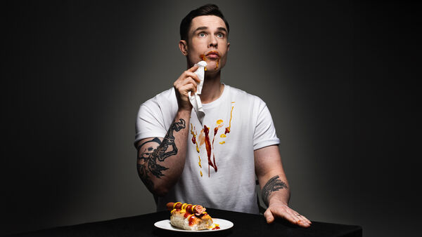 Ed Gamble eating a hotdog with ketchup and mustard spilled down his white t-shirt
