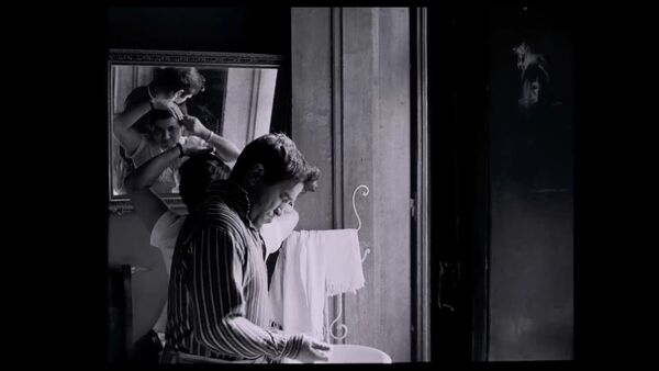 A black and white image of a man getting ready in a bathroom. A woman can be seen looking into a mirror in the background. A black and white image of a man getting ready in a bathroom. A woman can be seen looking into a mirror in the background.