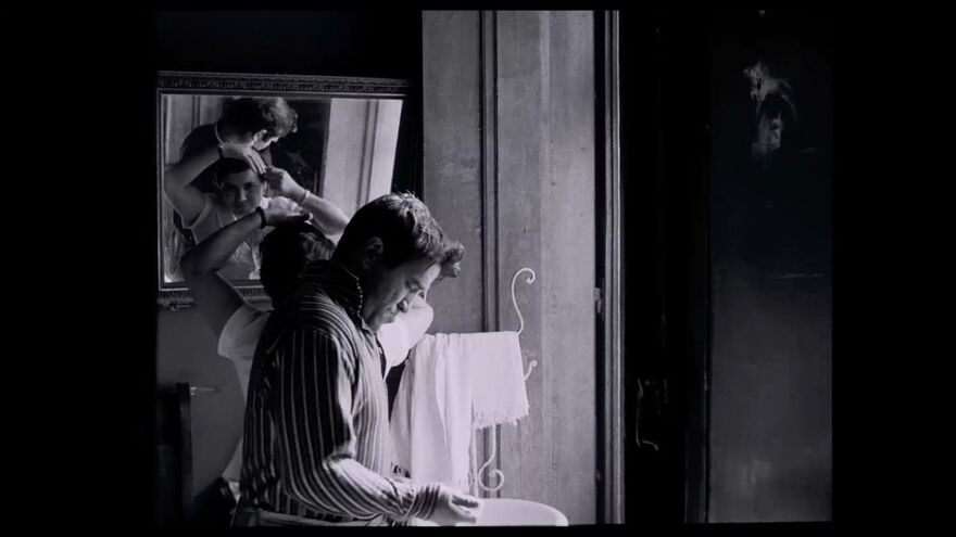 A black and white image of a man getting ready in a bathroom. A woman can be seen looking into a mirror in the background. 