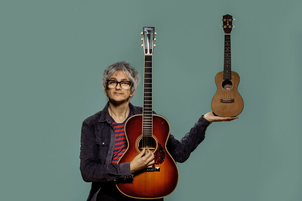 Nick cope holding a guitar and a Ukulele with a bluey green background