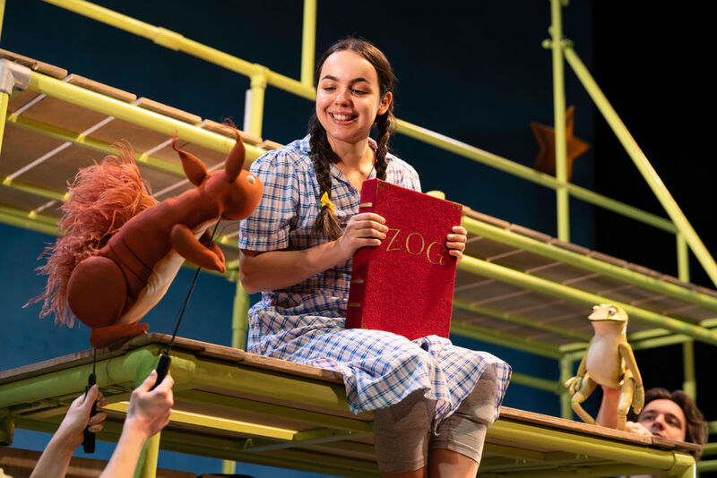 A smiling actor from the Zog theatre production sits on a stage set holding a large red Zog book, interacting with squirrel and frog puppets.
