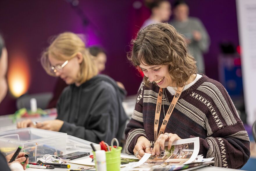 A smiling University of Warwick student making a paper collage at a busy arts and crafts table.