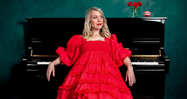 Rachel Parris in a long red dress leaning on a piano 