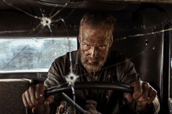 An older man behind the wheel of a car. Bullet holes can be seen in the windshield. 