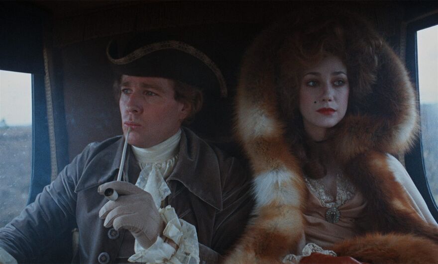A man and woman sit together in a carriage, both wearing period dress.