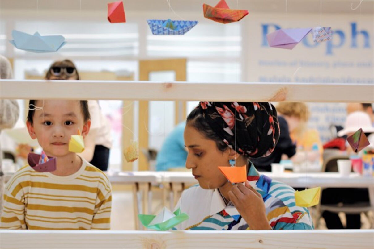 Mother and son attach origami to strings hanging from art work display