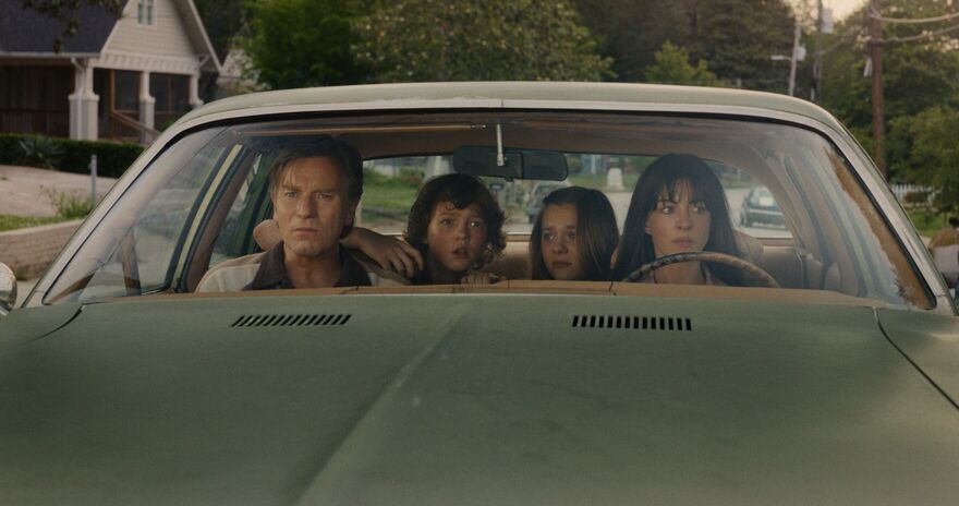 A family of four sit together looking nervous inside a green car. 