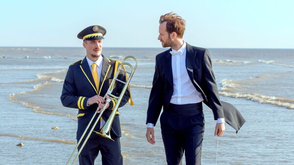 A conductor and trombone player, both smartly dressed, stand together on a beach. 