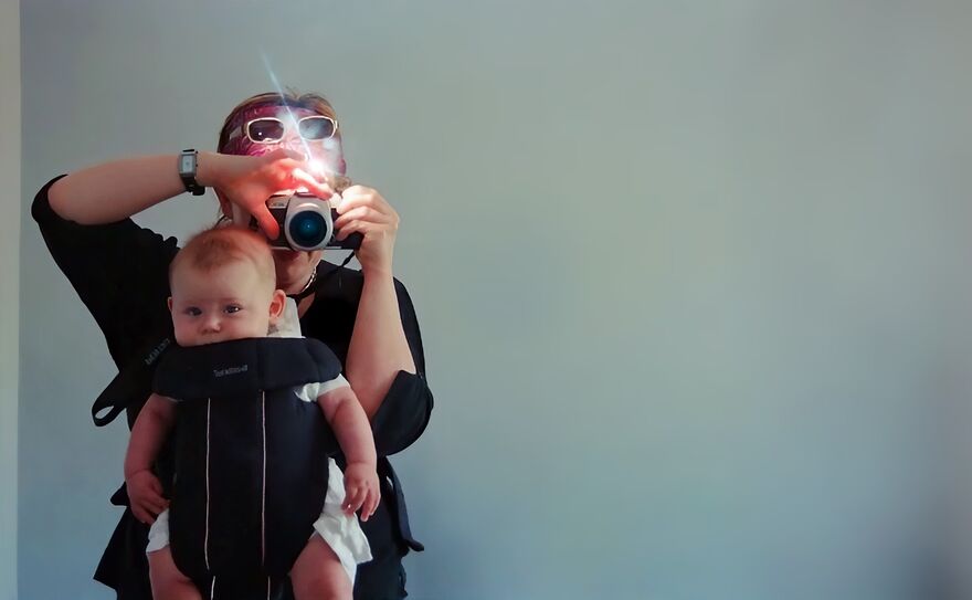 A woman takes a photograph as she holds a baby in a carrier on her front. 