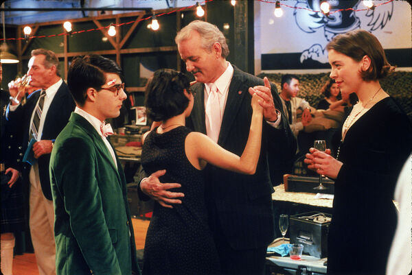 A young couple stand facing each other at a school dance. An older couple dances between them. 