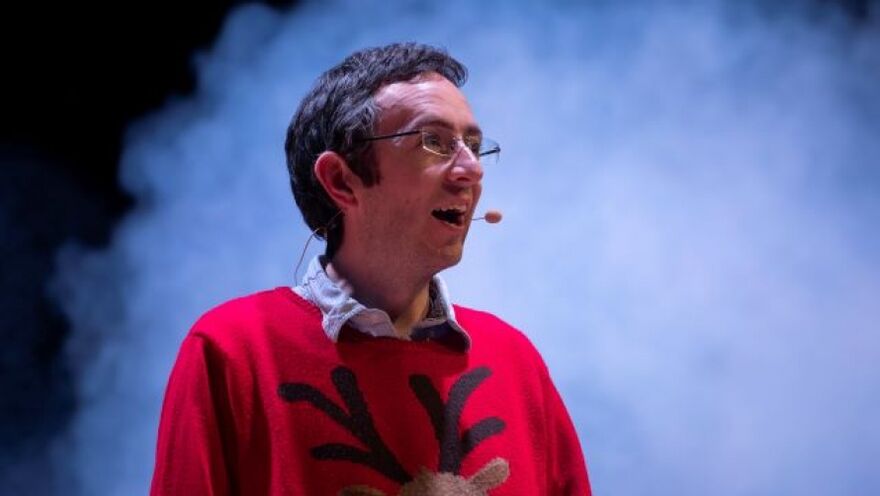 A man with a reindeer jumper on stands on a smoky stage